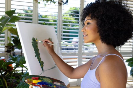 Biracial young woman painting on canvas at home, standing by window. She has curly black hair, wearing white tank top, holding a palette, unaltered