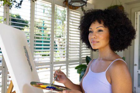 Biracial woman painting on canvas at home, wearing white tank top. She has curly black hair, light brown skin, and is holding a palette, unaltered