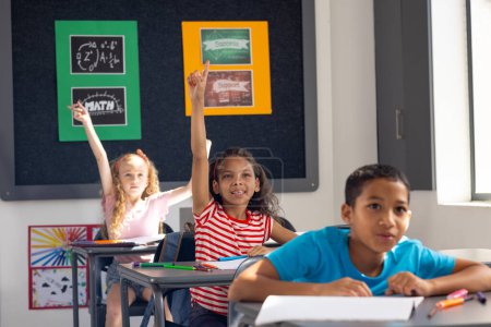 In school, diverse young students raising hands in the classroom, eager to answer. Caucasian girl with blonde hair, biracial boy with dark hair looking forward, both wearing casual clothes, unaltered