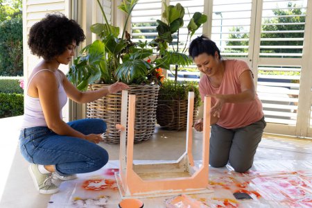 Biracial mother and adult daughter painting furniture together at home in an upcycling project. Both wearing casual clothes, mother has short black hair, daughter curly, unaltered