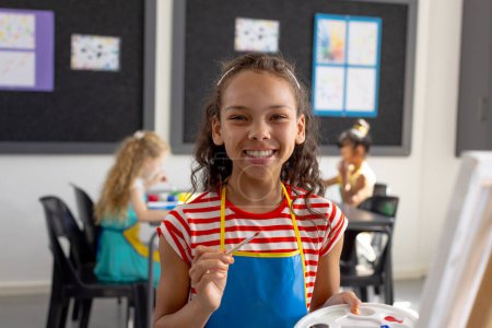 In school art class, biracial young girl holding a paintbrush smiles at camera. Behind her, diverse young friends focus on creating paintings, unaltered.