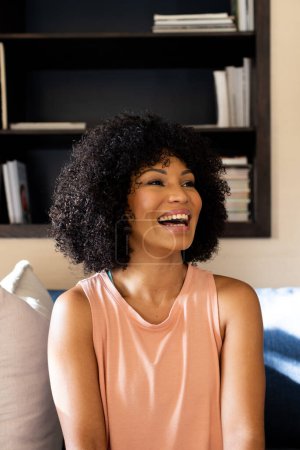 Biracial young woman laughing, sitting on couch at home. She has curly black hair, light brown skin, and wears a peach top, unaltered