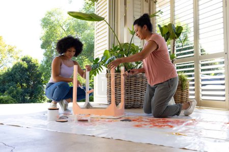 Biracial mother and adult daughter are painting furniture outside at home for an upcycling project. Mother has short black hair, daughter has curly brown hair, unaltered.