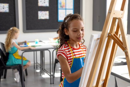 In school, during art class, a young biracial girl wearing a striped apron is painting. Behind her, a young Caucasian girl focuses on her own artwork, unaltered.