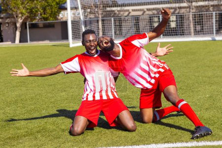 Zwei junge afroamerikanische männliche Athleten feiern auf einem Fußballplatz im Freien. Beide tragen rot-weiß gestreifte Trikots, sie strahlen Freude aus, unverändert.