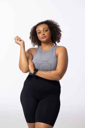 Biracial young female plus size model with curly brown hair standing confidently on white background. She has light brown skin, wearing a gray top and black shorts, unaltered, embodying body positivity.