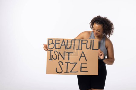 Biracial young female plus size model holds poster on a white background. She has curly hair, a fit build, wears a tank top and leggings, and promotes body positivity, unaltered