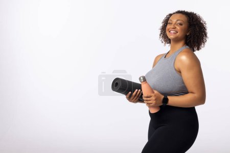 Biracial young female plus size model holds yoga mat and water bottle, copy space. She smiles against a white background, her curly brown hair and light brown skin complementing her sportswear