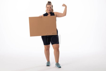 A Caucasian plus-size model with white background holds a sign, copy space. She sports blonde hair, a black top, and shorts, exuding confidence and promoting body positivity, unaltered