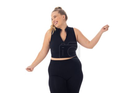 Caucasian young female plus size model on white background wearing black activewear, dancing, copy space. She has blonde hair, blue eyes, and is young, unaltered, embodying body positivity.