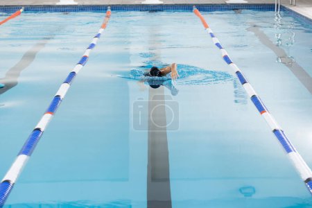 Biracial girl in goggles swims freestyle in indoor pool lane, copy space. Water splashing around as she moves swiftly through clear blue water, unaltered