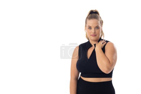 Plus size Caucasian young female model in black sportswear on white background, copy space. She has blonde hair, blue eyes, and is touching her shoulder, embodying body positivity, unaltered