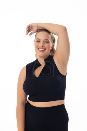 Smiling Caucasian plus-size model poses, hand on head, against white background. She has blonde hair, blue eyes, and wears black outfit, embodying body positivity, unaltered