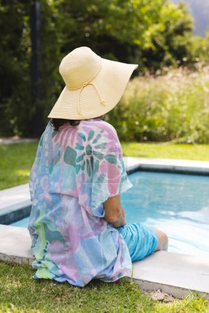 Outdoors, Caucasian senior woman wearing a hat, relaxing by pool. Sporting brown hair, donning a colorful shawl and blue pants, with a serene blue background, unaltered