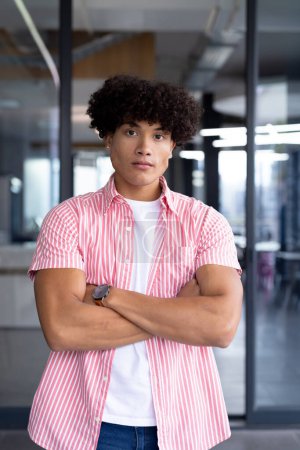 Dans un bureau moderne, un jeune travailleur masculin biracial debout avec les bras croisés. Cheveux bruns bouclés et peau marron clair, vêtu d'une chemise à rayures rouges, il respire la confiance, intacte
