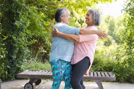 Outdoors, diverse senior female friends embracing warmly. Sporting short gray hair and wearing casual clothes, they're sharing smiles, enjoying sunny day with a green background, unaltered