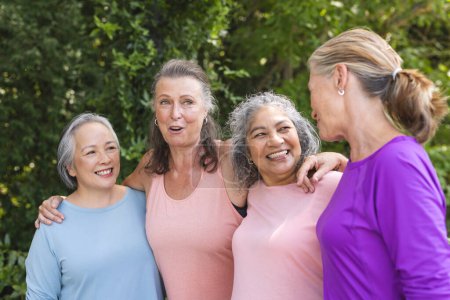 Outdoors, diverse senior female friends standing together, smiling. Wearing casual clothes with visible wrinkles, they're enjoying serene green background, relishing moment, unaltered