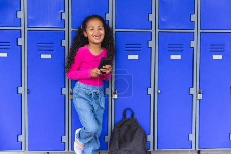 In school beside lockers, a biracial young girl is using a smartphone. She has curly dark hair, wearing a pink top and blue jeans, unaltered.