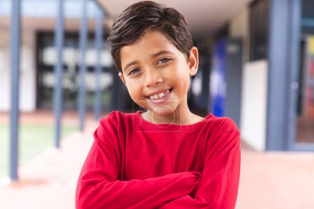 In der Schule, draußen, ein kleiner Junge mit zwei Rassen, der in die Kamera lächelt. Er hat kurze dunkle Haare, hellbraune Haut und trägt ein rotes Hemd,