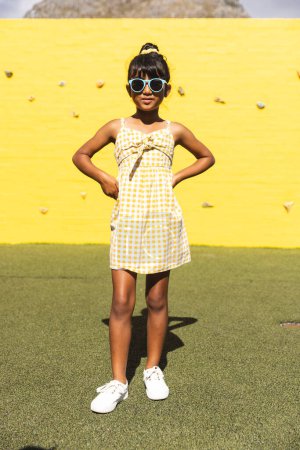 Zweirassiges Mädchen mit Sonnenbrille und Sonnenbrille posiert selbstbewusst im Freien. Ihre stylische Sommerkleidung suggeriert einen lustigen Tag in der Sonne.