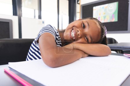 In school, young biracial girl with a bright smile is resting her head on her arms in the classroom. She has light brown skin, dark brown eyes, and her hair is styled in neat braids, unaltered.