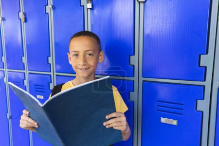 Biracial boy stands in front of school lockers, with copy space. He smiles while holding a book, capturing a moment of school life.