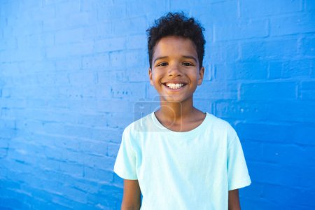 Biracial boy smiles brightly against a blue wall. His cheerful expression adds a lively touch to the outdoor setting.