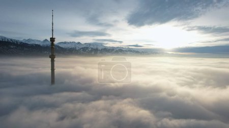 Photo for The TV tower looks out of the clouds at sunset. Top view from a drone on a double layer of clouds. Yellow-orange color from the rays of the sun. Shadows on clouds. High mountains in the distance - Royalty Free Image