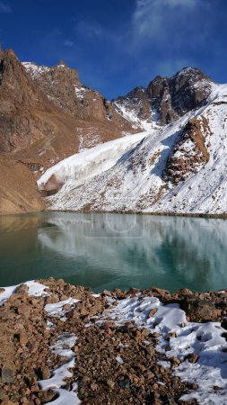 Ein Bergsee mit smaragdgrünem Wasser spiegelt einen Gletscher wie einen Spiegel wider. Man sieht die Gipfel der Berge. Der See ist teilweise zugefroren. Stellenweise liegen große Steine und Schnee. Moränensee