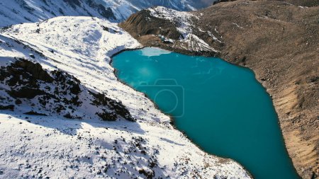Foto de A mountain lake with blue water looks like a mirror. Top view from a drone. The moraine lake is partially frozen. In places there are large rocks and snow, a glacier. Pipes and a house are visible - Imagen libre de derechos