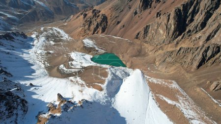 Foto de A mountain lake with emerald water is like a mirror. Top view from a drone. The moraine lake is partially frozen. There are large stones and snow in places, a glacier. Pipes and a house are visible - Imagen libre de derechos