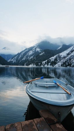 Ein Bergsee im Wald mit verspiegeltem Wasser. Holzsteg mit weißen Booten. Das Wasser spiegelt die Landschaft eines wolkenverhangenen Himmels wider, schneebedeckte Berge und Gipfel, Nadelhölzer. Kolsai-See, Kasachstan