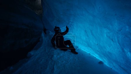 Photo for The climber made a halt inside an ice cave. The turquoise color of the ice gives a special atmosphere. The guy is looking for something in his backpack. There is snow on the icy floor. In mountains - Royalty Free Image