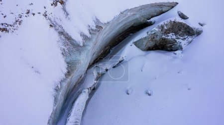 Foto de A huge ice wall from a glacier in the mountains. Everything is covered with snow and large stones. Top view from the drone on the cracks in the ice. The glacier breaks, the ice flakes off. Almaty - Imagen libre de derechos
