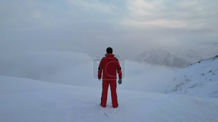 Photo for A climber in a red suit stands at the top of the mountain. Looks at the heavenly ocean of white clouds. There is snow all around, blue sky, dawn and calm. An epic moment of life. There are tours - Royalty Free Image