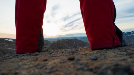 Téléchargez les photos : Un type en bottes de trekking fait des pas à travers le Canyon. Il s'arrête au bord. Au lever du soleil. Rayons orange-rouge du soleil et nuages bleus. La terre du canyon rouge. De petites pierres. Pantalon rouge sport. La vue est comme sur Mars - en image libre de droit