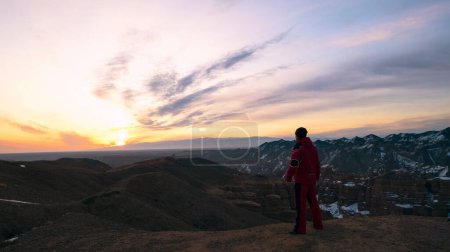 Téléchargez les photos : Le gars en rouge marche jusqu'au bord du canyon. Lumière rouge-orange du soleil levant. Nuages violets. Canyon orange Charyn. Bonjour. Il y a de la neige par endroits. Un voyage à travers le canyon. Kazakhstan - en image libre de droit