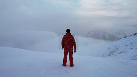 Photo for A climber in a red suit stands at the top of the mountain. Looks at the heavenly ocean of white clouds. There is snow all around, blue sky, dawn and calm. An epic moment of life. There are tours - Royalty Free Image
