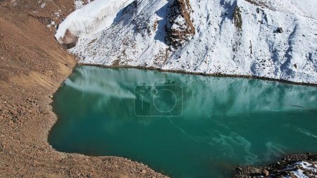 Foto de Un lago de montaña con agua esmeralda refleja un glaciar como un espejo. Se pueden ver los picos de las montañas. El lago está parcialmente congelado. Hay grandes piedras y nieve en algunos lugares. Lago Moraine - Imagen libre de derechos