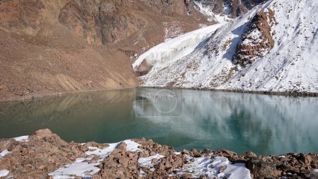 Foto de Un lago de montaña con agua esmeralda refleja un glaciar como un espejo. Se pueden ver los picos de las montañas. El lago está parcialmente congelado. Hay grandes piedras y nieve en algunos lugares. Lago Moraine - Imagen libre de derechos