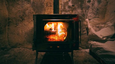 Photo for A fire is burning in a camping stove with a glass door. The process of smoldering fuel briquettes is visible. The lining of the stove made of mirror metal reflects the gray old walls of the hut. - Royalty Free Image
