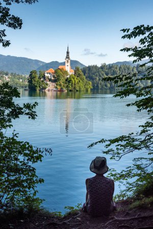 Paysage de Slovénie. La silhouette d'une personne assise se distingue des eaux bleues du lac de Bled. En arrière-plan, on peut voir l "Église de la Mère de Dieu, aussi appelée Église de pèlerinage de l'Assomption de Marie