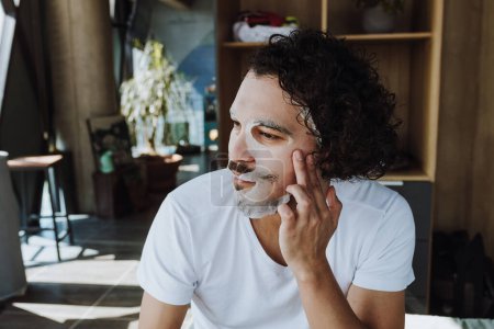 Photo for Hispanic Man applying exfoliating facial mask treatment taking care of skin, morning face wash routine for cleaning in loft apartment in Mexico Latin America - Royalty Free Image