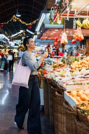 Foto de Mujer joven hispana con cabeza rapada o pelo corto comprando verduras en el mercado tradicional o en la tienda de comestibles - Imagen libre de derechos