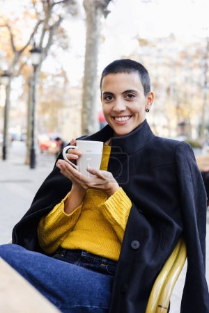 Foto de Mujer hispana joven skinhead beber taza de café en la ciudad urbana cafetería terraza o cafetería, Retrato de mujer latina con pelo corto en la calle al aire libre en la temporada de otoño - Imagen libre de derechos