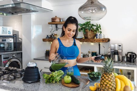 Foto de Mujer latina vertiendo jugo verde de la batidora en el vidrio en la cocina en casa en México América Latina, mujer hispana en la dieta de desintoxicación - Imagen libre de derechos