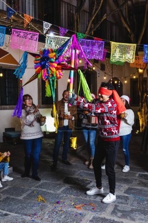 Photo for Latin young man breaking a pinata at traditional mexican posada celebration for Christmas eve in Mexico Latin America, hispanic family in holidays - Royalty Free Image