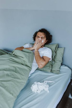 Kranker lateinamerikanischer Mann pustet sich die Nase aus, während er zu Hause in Mexiko im Bett liegt