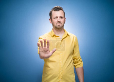 Mann gestikuliert Stoppschild mit Handfläche, weigert sich oder lehnt etwas vor blauem Hintergrund ab, trägt gelbes Hemd. Studioaufnahme