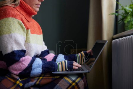 Foto de Mujer en guantes con el ordenador portátil tratando de mantener caliente por el radiador durante el costo de vida crisis de energía - Imagen libre de derechos